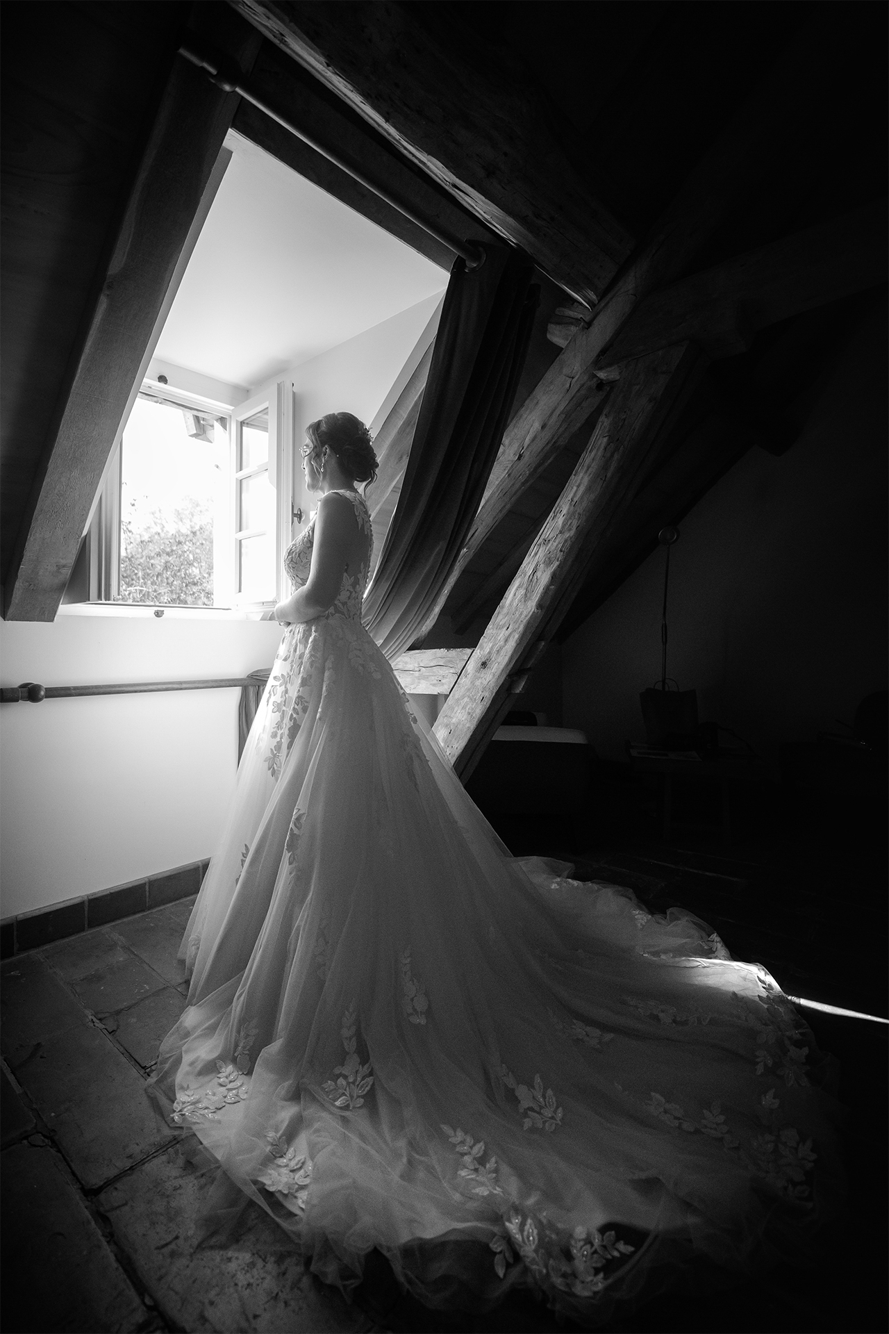 La mariée près d’une fenêtre dans une pièce mansardée pendant ses préparatifs, en noir et blanc.